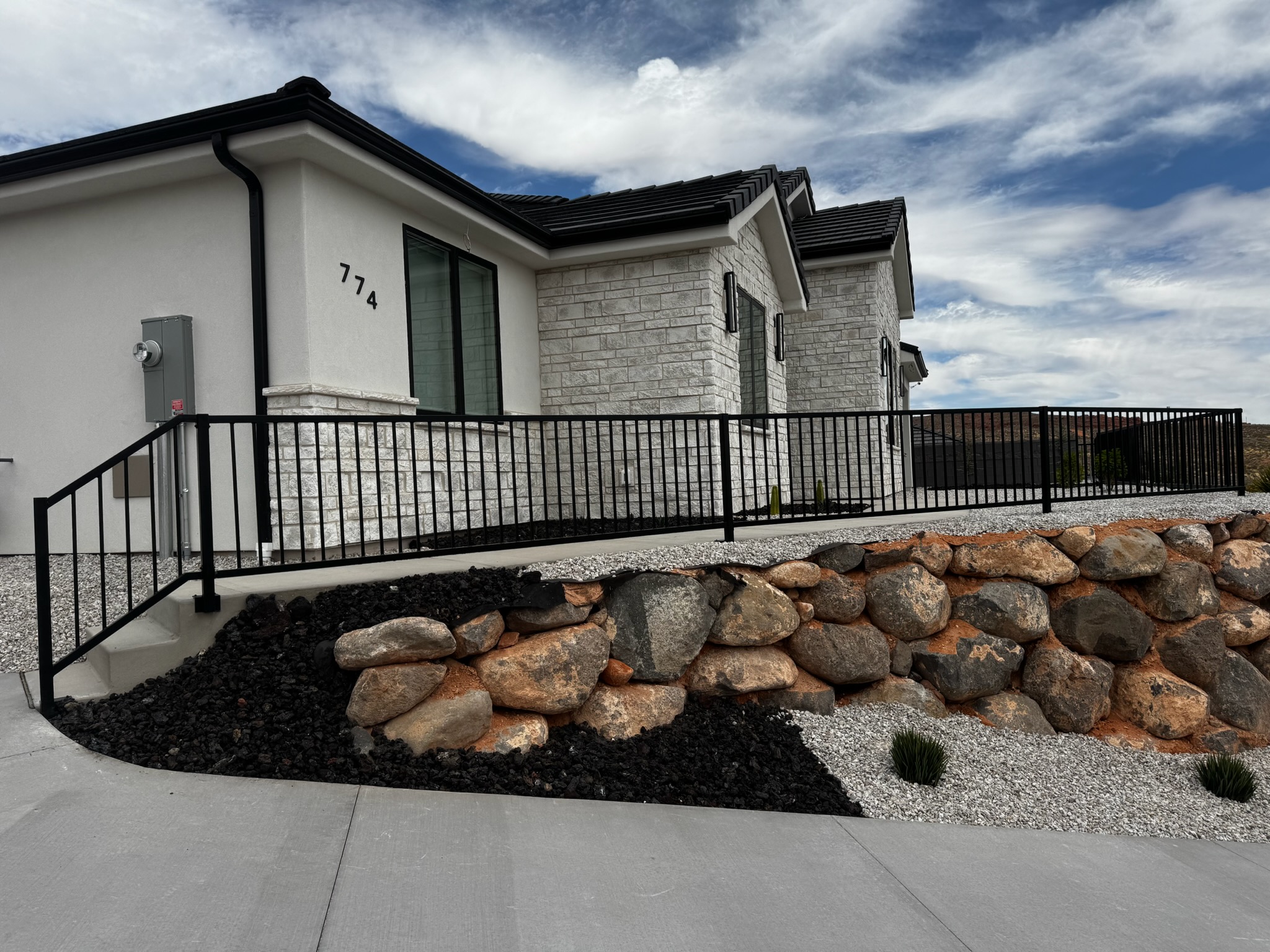 Black steel walkway railing with vertical pickets along a concrete path and entry steps above a natural boulder retaining wall in St. George, Utah