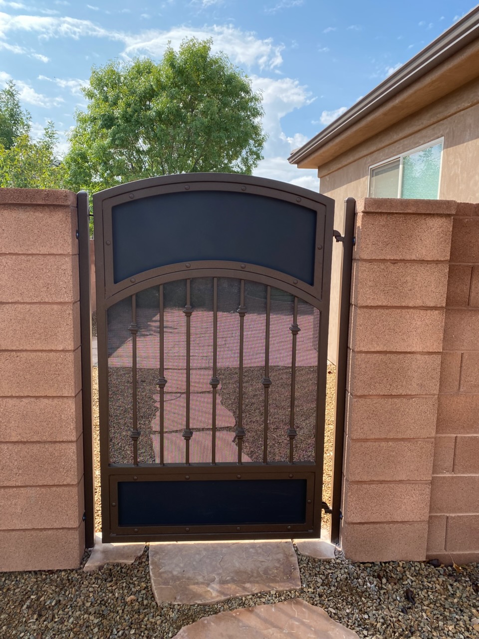 Arched bronze steel side-yard gate with decorative knuckle pickets, mesh screen, and rivet accents between block wall pillars in St. George, Utah