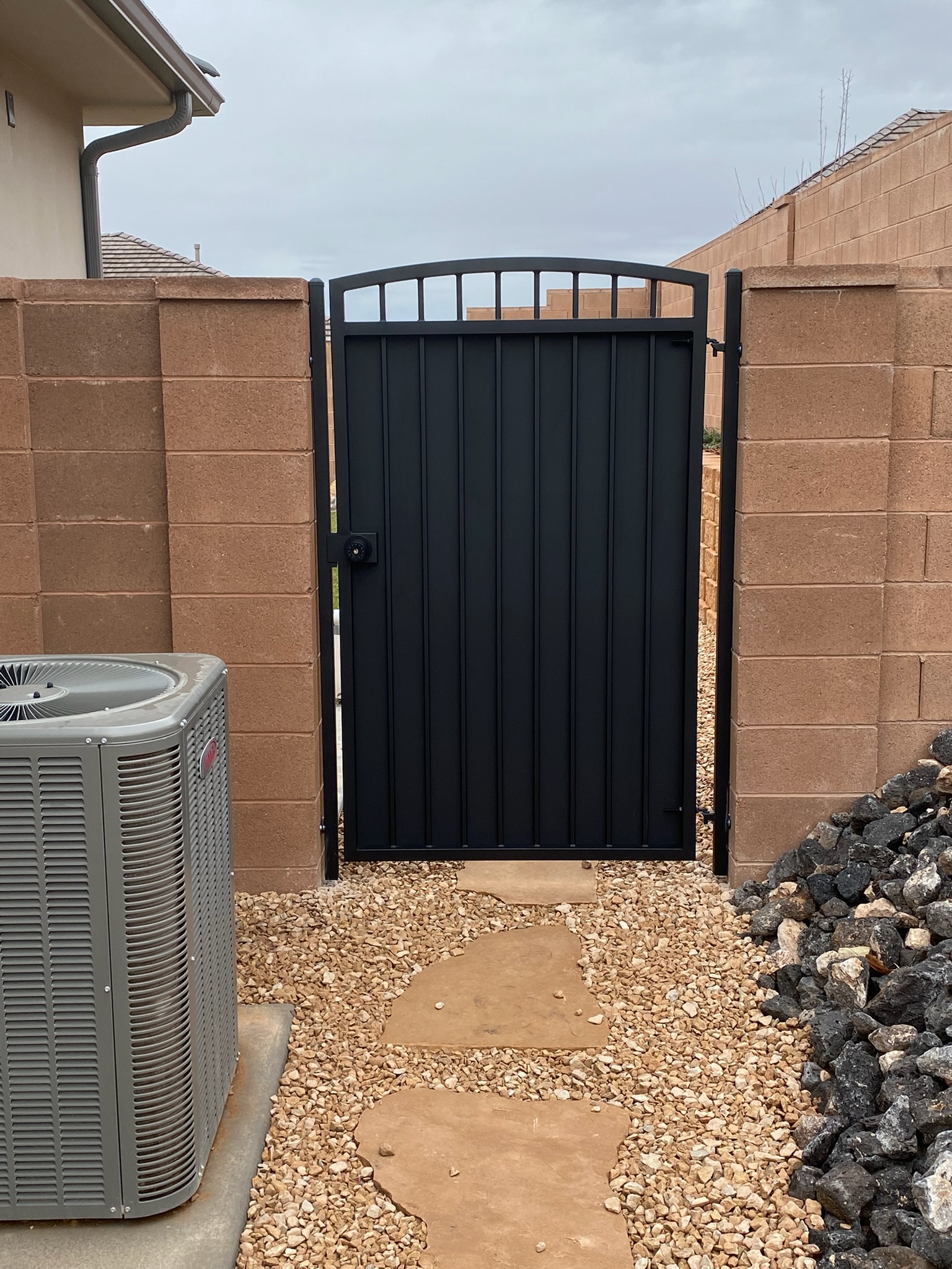 Black steel side-yard gate with arched picket top, vertical ribbed privacy panel, and keypad lock between block wall pillars in St. George, Utah