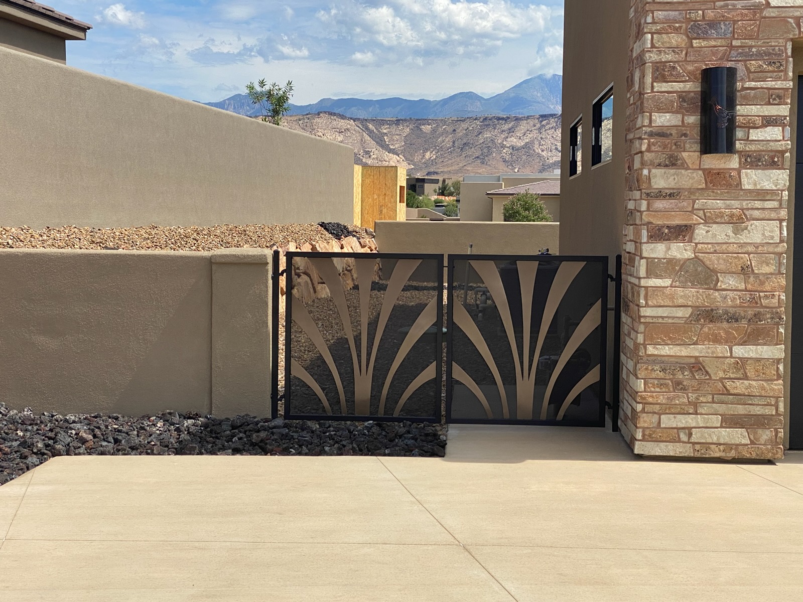 Black steel double gate with decorative cut-metal panels featuring a bronze fan-shaped design, installed between a stucco wall and stone veneer column in St. George, Utah