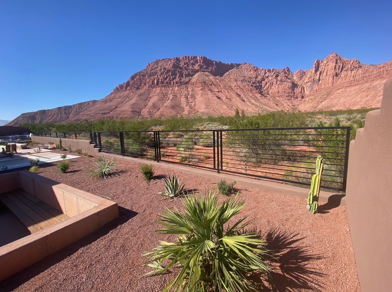 Black steel horizontal bar view fence mounted on a low stucco wall around a desert-landscaped backyard with red rock cliffs in the background in St. George, Utah