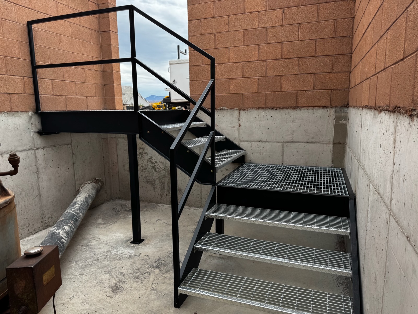 Black steel access staircase with galvanized bar grating treads, diamond plate nosing, a grated landing, and handrails leading into a below-grade concrete pump room in St. George, Utah