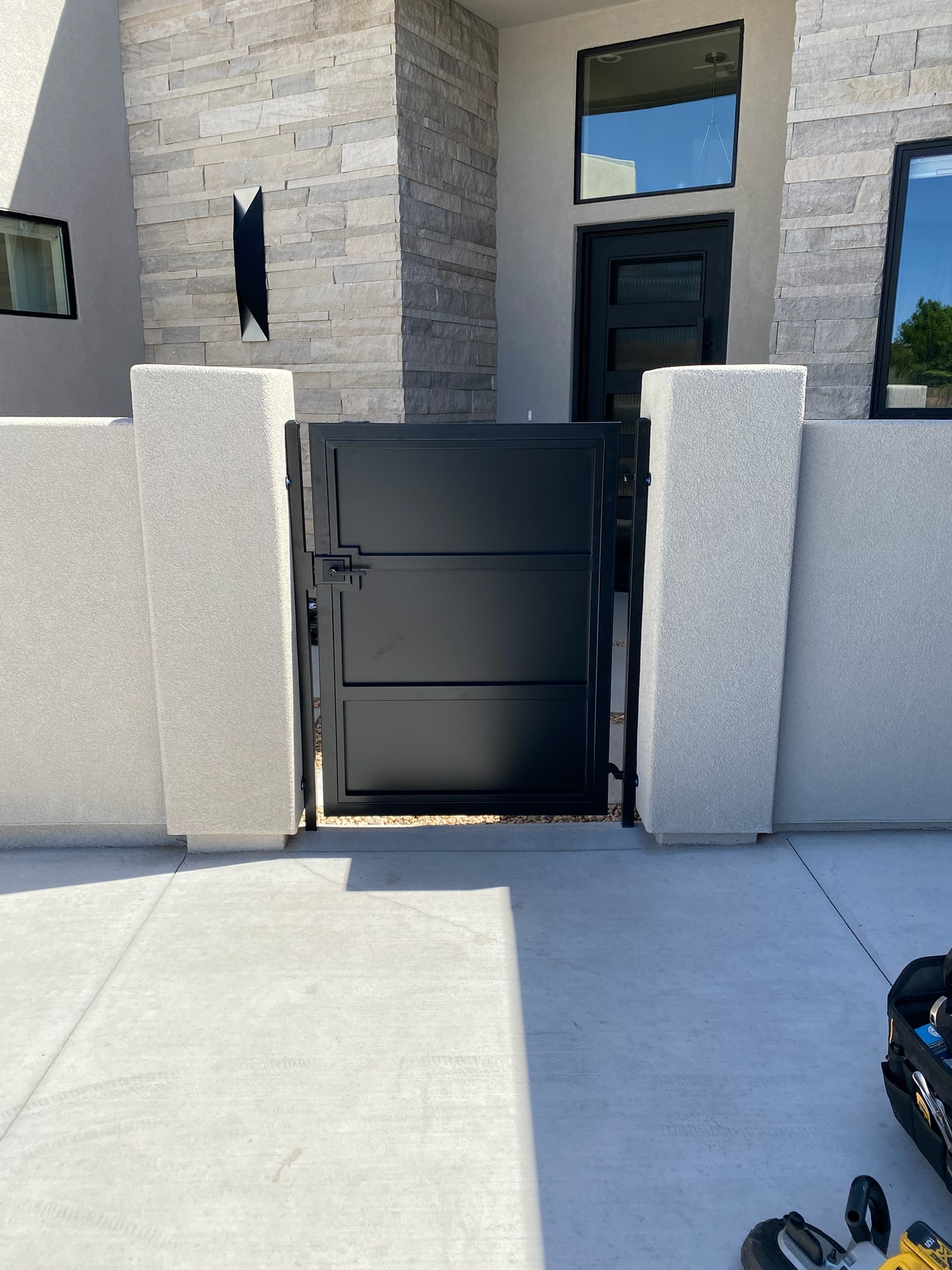 Modern solid black steel front entry courtyard gate with three horizontal panels and a square lever latch between light stucco pillars in St. George, Utah