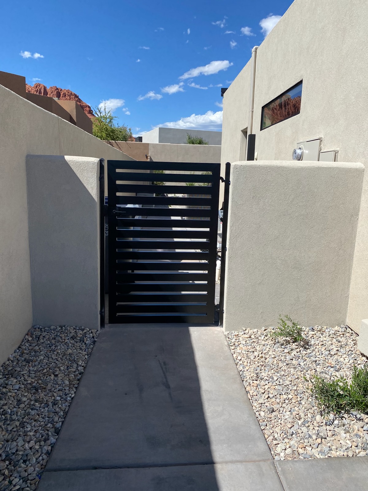 Modern black steel walk-through gate with wide horizontal slats installed between smooth stucco wall pillars at a contemporary desert home in St. George, Utah