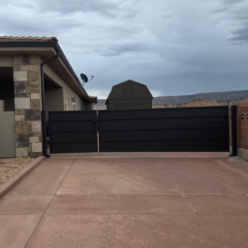 Black steel unequal double swing RV gate with horizontal panel sections spanning a wide concrete driveway between a stucco home and block wall, with red rock mesas behind, in Southern Utah